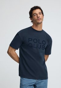 Marinblå kortärmad t-shirt med "POLO CLUB" tryckt i en något mörkare nyans. Rundad hals och avslappnad passform. Bärs med blå jeans.