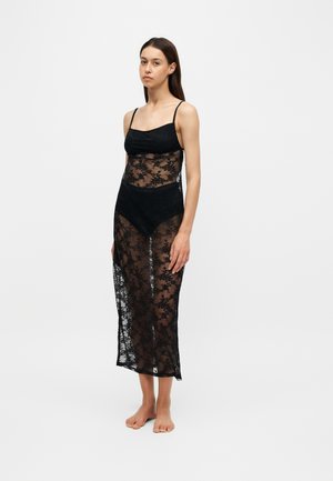 Femeie stând desculță purtând o rochie neagră de dantelă transparentă cu modele florale peste lenjerie neagră, pe un fundal simplu, deschis.