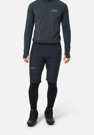 Homme portant un haut noir à manches longues zippé, un short noir matelassé, des leggings noirs et des bottes noires à fermeture éclair, debout devant un fond blanc.