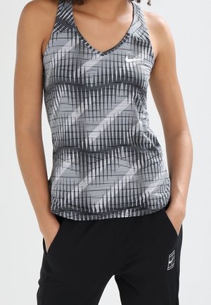 Femme portant un haut de sport sans manches à motifs noir, blanc et gris et un pantalon noir, les mains dans les poches.