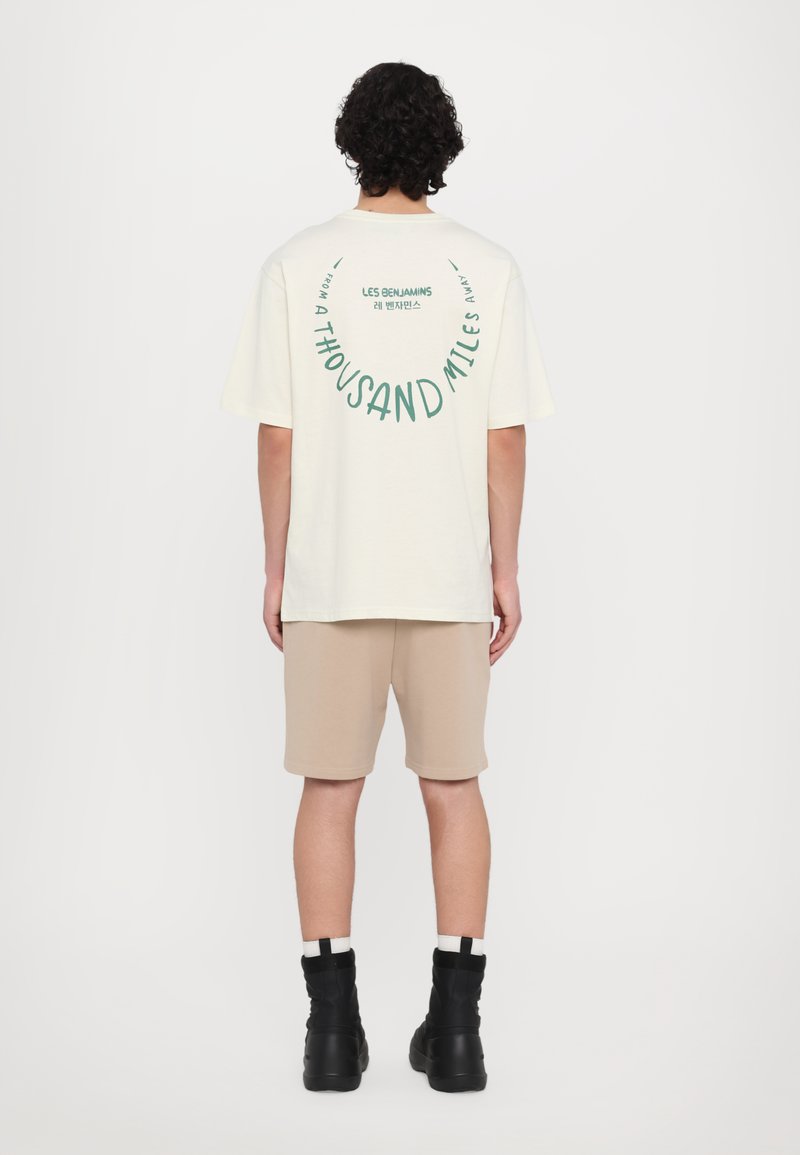 T-shirt crème à manches courtes avec un texte graphique vert à l'arrière. Porté avec un short beige et des bottes montantes noires. Design minimaliste.