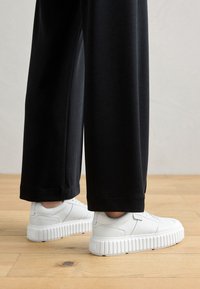 Des baskets blanches avec une semelle texturée et épaisse, assorties à un pantalon noir ample. Les chaussures présentent un cuir lisse et des éléments de design minimalistes.