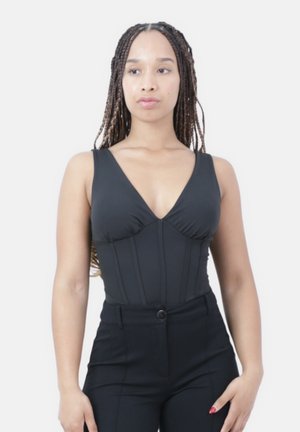 Mujer con largas trenzas de pie de frente, vestida con un top negro sin mangas de escote en V y pantalones negros sobre un fondo blanco liso.