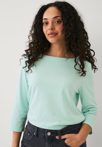 Top lungo maniche verde menta con scollatura a barca, in morbido cotone e vestibilità rilassata, abbinato a jeans scuri con texture.