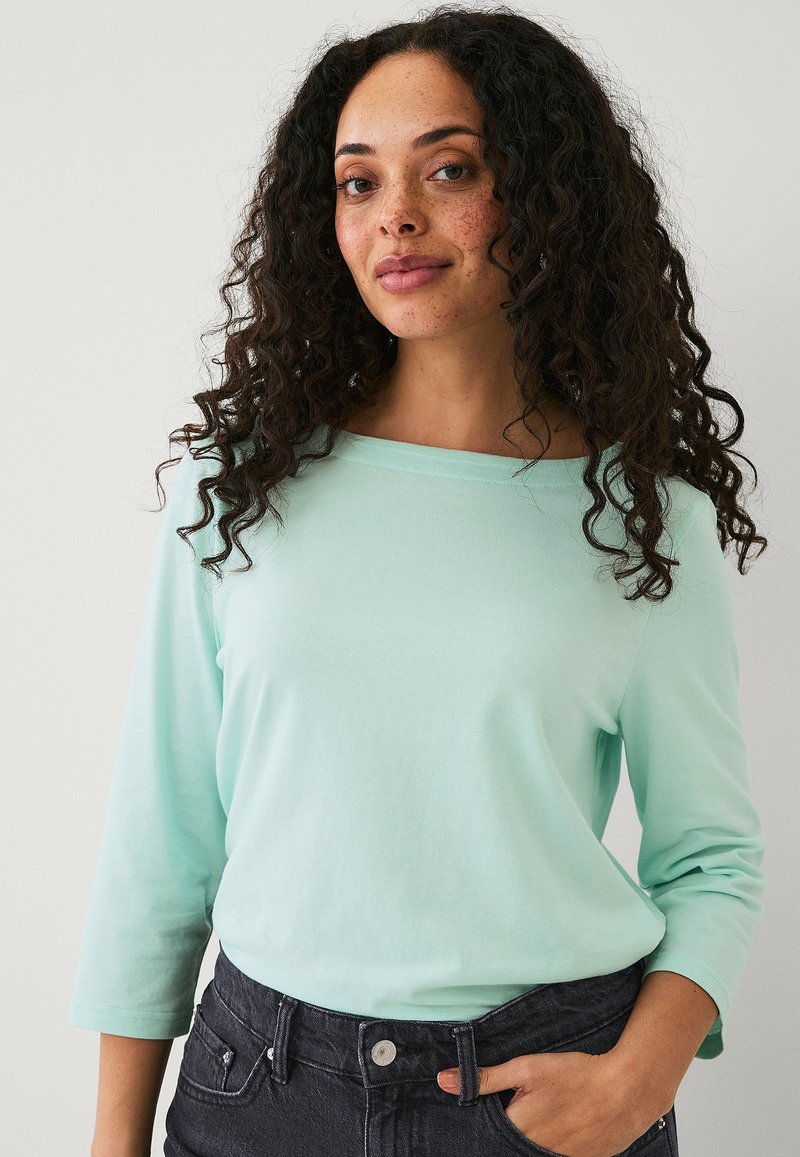 Top lungo maniche verde menta con scollatura a barca, in morbido cotone e vestibilità rilassata, abbinato a jeans scuri con texture.