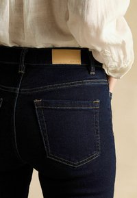 Personne portant un jean en denim bleu foncé avec des coutures visibles sur la poche arrière et une étiquette de marque en cuir marron clair, associé à une chemise blanche à manches longues.