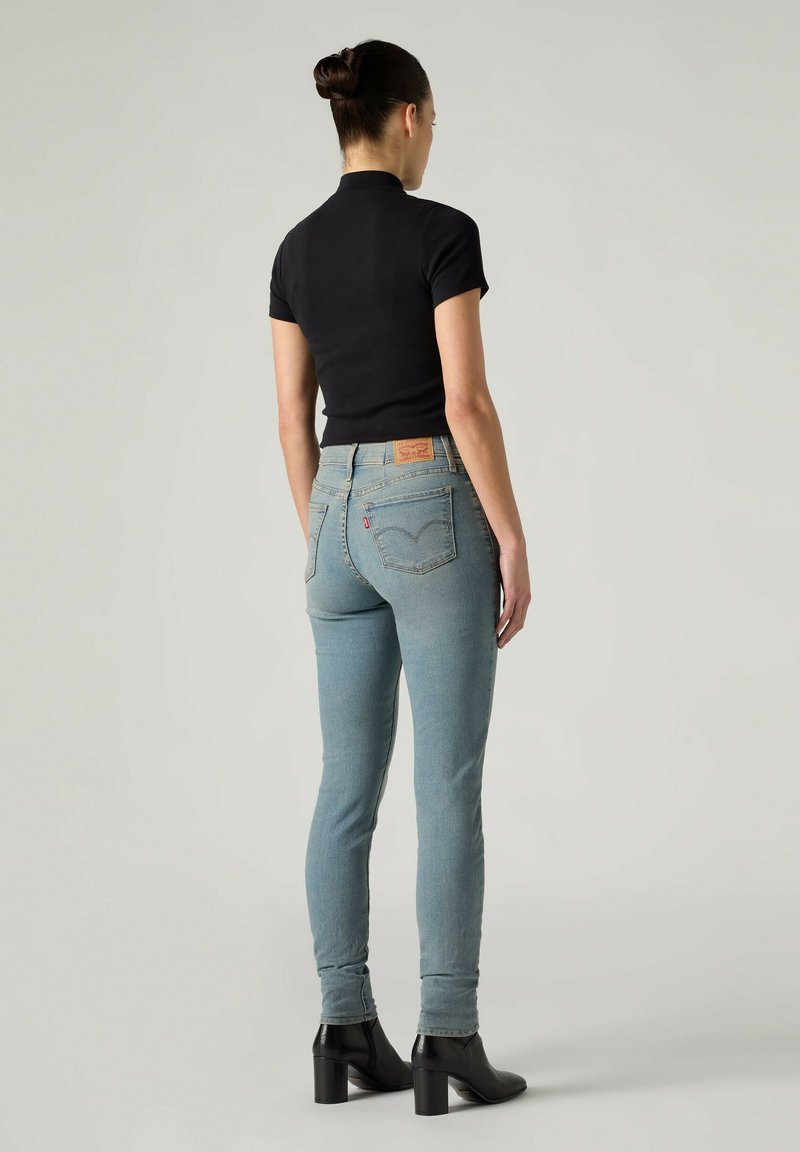 Kvinde med knold, iført sort kortærmet top, lyseblå højtaljede jeans og sorte ankelstøvler med hæl, stående mod en ensfarvet baggrund.
