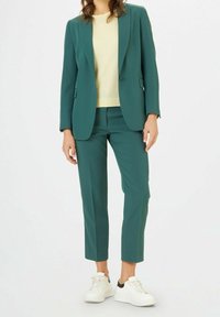 Costume teal avec blazer ajusté et pantalons courts, associé à un pull jaune clair et des baskets blanches avec des accents noirs.