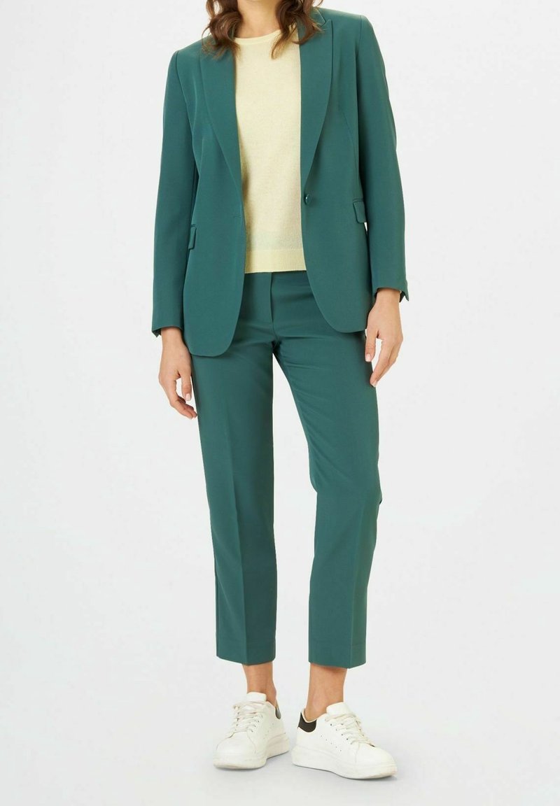 Costume teal avec blazer ajusté et pantalons courts, associé à un pull jaune clair et des baskets blanches avec des accents noirs.