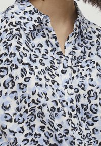 Primo piano di una persona che indossa una camicia abbottonata con un motivo a stampa leopardata blu e nera e colletto aperto.