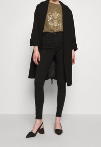 Abrigo negro sobre una camiseta gráfica marrón con un diseño dorado, combinado con jeans ajustados negros y zapatos de tacón negro con tira trasera.