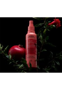 Après-shampooing sans rinçage Aveda Nutri Plenish dans un flacon vaporisateur rose vif, avec une grenade arrondie et des feuilles vertes sur un fond noir.