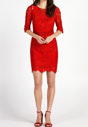 Kvinde iført knælængde rød blondekjole med blomsterprint og trekvartlange ærmer, kombineret med røde højhælede sandaler med ankelrem.