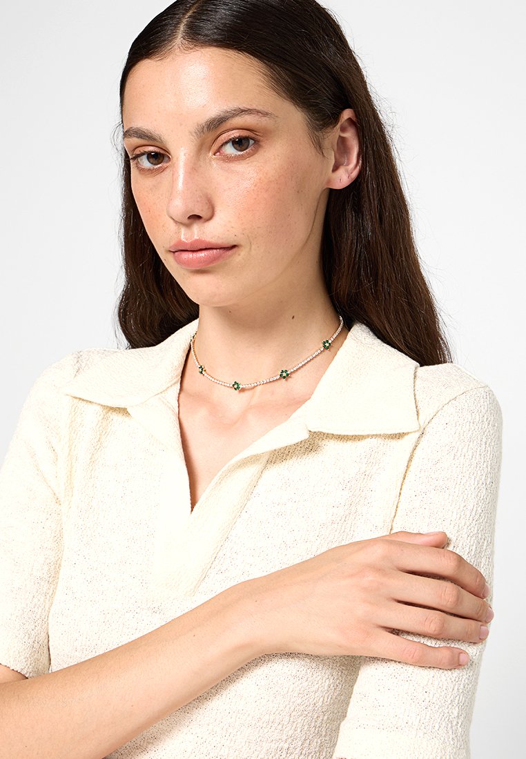 Collana d'oro con piccoli dettagli a forma di fiore verde e perline trasparenti, di fronte a una persona che indossa una polo di colore crema con una texture particolare.