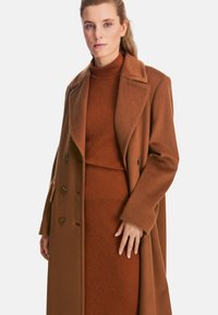 Brown wool blend coat with a double-breasted design, large lapels, and buttons. Layered over a matching ribbed turtleneck dress with subtle patterns.
