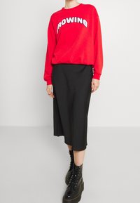 Czerwona bluza z napisem "BROWNING" w białym kolorze, zestawiona z czarną spódnicą midi i czarnymi sznurowanymi botkami. Gładka tkanina, luźny krój.