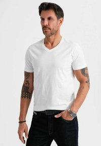 Camiseta blanca de cuello en V hecha de algodón, de mangas cortas, con textura suave, combinada con jeans oscuros y un reloj, con tatuajes visibles en el brazo.