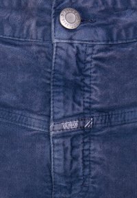 Dunkelblaue Denim-Stoff mit feiner Textur, ausgestattet mit einem runden Metallknopf und einem Markenetikett mit der Aufschrift "CLOSED". Sichtbare Nähte.
