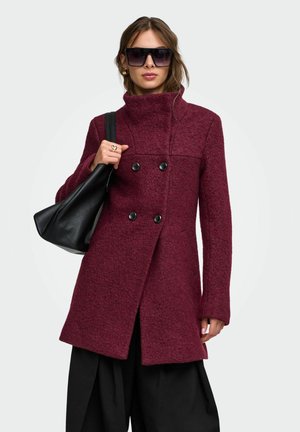 Femme portant de grandes lunettes de soleil noires et un manteau bordeaux à double boutonnage, tenant un sac à main noir sur l'épaule.