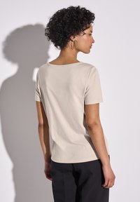 T-shirt beige à manches courtes avec un col ras du cou, texture lisse et coupe ajustée, associée à un pantalon sombre, vue de dos.