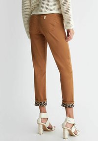 Kobieta w brązowych spodni w mankietach z podszewką w leopardzie plamki oraz białych sandałach na obcasie, stojąca na jednolitym jasnym tle.