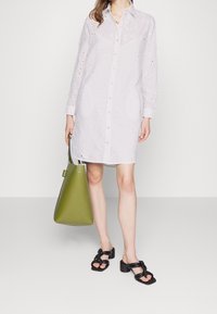 Vestido camisero de algodón blanco con patrón de ojales, cierre frontal de botones, mangas largas y cuello. Combinado con un bolso tote verde y sandalias negras.