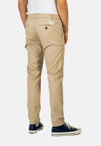 Chinos color caqui con un corte slim, que incluyen un bolsillo trasero con cierre de botón y están combinados con zapatillas azules. Tejido de algodón suave, diseño casual.