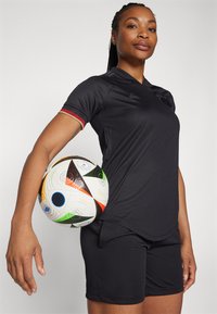 Svart idrottströja med korta ärmar, med en subtil struktur och färgglad kant. En flerfärgad fotboll hålls under armen.