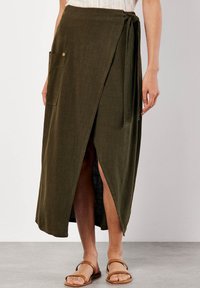 Olivgrüne Wickelrock aus einem strukturierten Stoff mit einem Schlitz vorne, einer Tasche und einem Knoten in der Taille. Kombiniert mit braunen Sandalen.