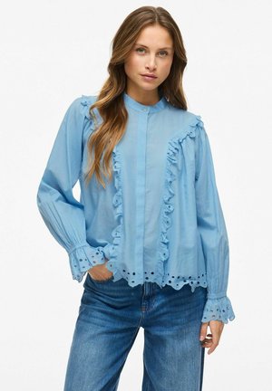 Lyseblå bluse med flæser langs fronten og ærmerne, med broderi ved kanten og rundt om manchetterne; knaplukning.