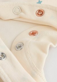Kurzarm-Overall in Beige mit metallischen Druckknöpfen in Silber-, Gold- und Kupferausführung, mit einer weichen Textur und dezenten blauen Sternen.