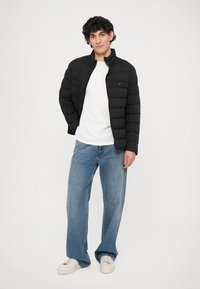 Zwarte pufferjas met horizontale naden, gedragen over een wit shirt, gecombineerd met lichtblauwe wijde jeans en witte sneakers.