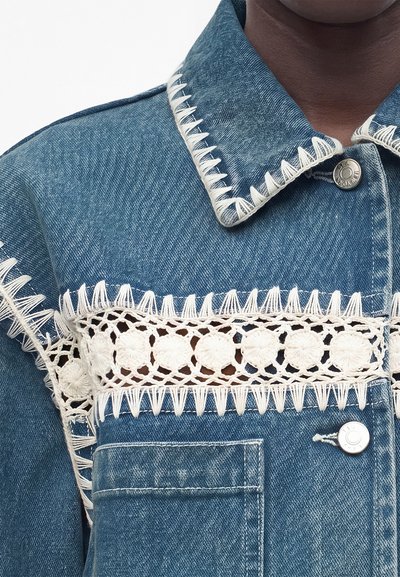 Κοντινό πλάνο μπλε τζιν μπουφάν με λευκή δαντέλα κροσέ στο στήθος και λευκές ραφές στο γιακά και τις ραφές.