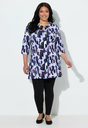 Kvinde iført et mønstret lilla tunika med trekvartærmer, sorte leggings og sorte flade sko, stående og smilende mod en ensfarvet baggrund.
