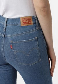 Ljusblå denimjeans med en rektangulär läderetikett vid midjan med röd text, samt bakfickor med distinkta sömnadsmönster.