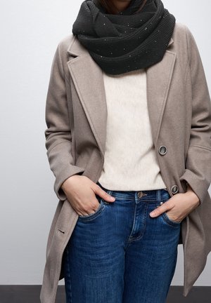 Personne portant un manteau beige, un pull blanc, un jean bleu et une écharpe en tricot noire, les mains dans les poches du jean, debout devant un fond uni.