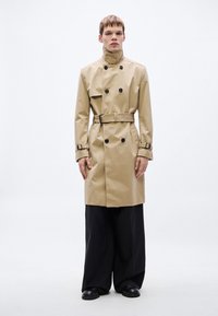 Béžový trenčkot s dvojradovým zapínaním, čiernymi gombíkmi, opaskom a nastaviteľnými manžetami, v kombinácii so širokými čiernymi nohavicami a čiernymi topánkami.