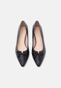 Paire de ballerines en cuir noir à bout pointu avec petits détails de nœuds sur fond blanc.