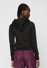 Sudadera de algodón negra con dobladillo y puños acanalados, que presenta una gran capucha forrada y mangas raglán. Combinada con pantalones morados.