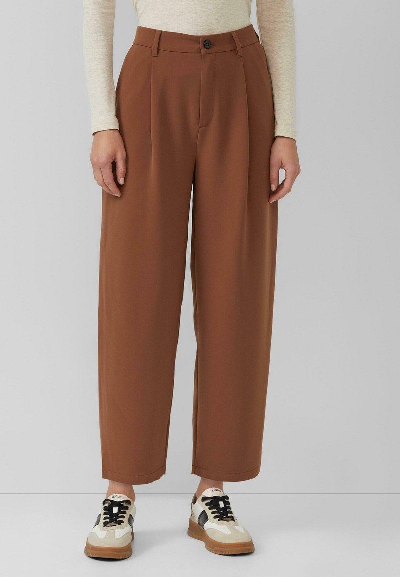 Brązowe spodnie o szerokich nogawkach wykonane z miękkiej tkaniny, z przodu marszczenia, zapinane na guzik, o gładkiej fakturze, zestawione z beżowymi trampkami.