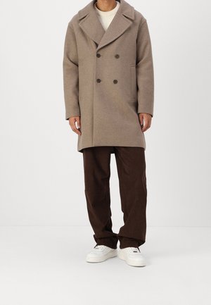 Hombre con abrigo de lana beige de doble botonadura, suéter blanco, pantalones de pana marrones y zapatillas blancas, de pie frente a un fondo sencillo.