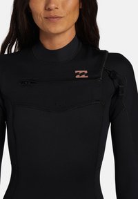 Muta nera con collo alto, dotata di zip frontale e chiusura laterale. Accentuata da un piccolo logo a onda rosa sul petto.