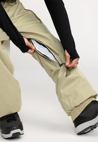 Beige ritssluitende broek met een meshvoering, voorzien van een verstelbare boeim en een relaxte pasvorm. Zwarte schoenen met een zichtbare gripsole.