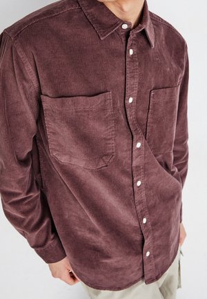 Homme portant une chemise en velours côtelé marron à manches longues avec des boutons devant et deux poches poitrine, debout devant un fond uni.