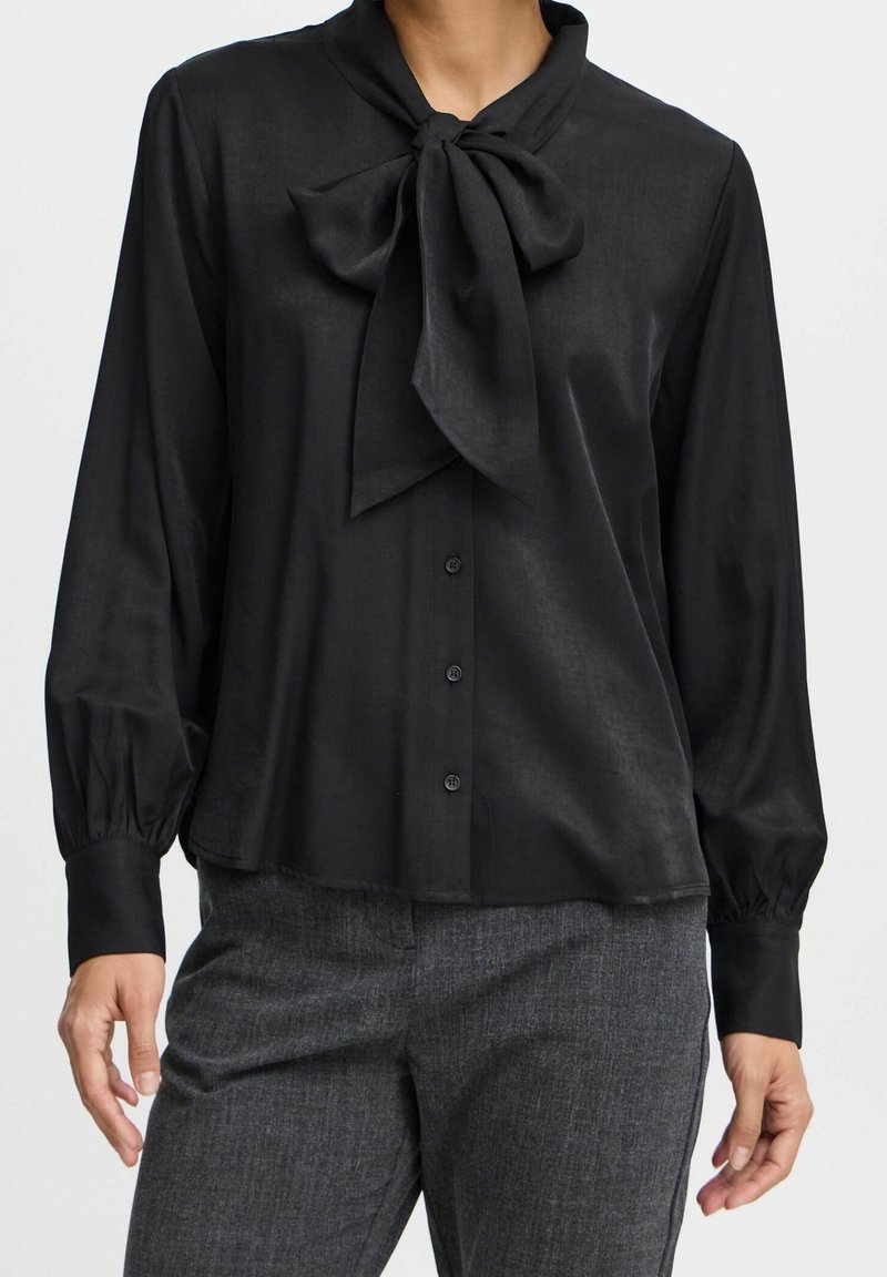 Femme portant un chemisier noir à manches longues avec un grand nœud papillon au col et un pantalon gris sur mesure, debout les mains détendues.