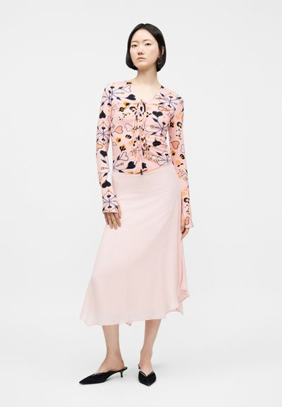 Γυναίκα που φοράει ροζ floral εφαρμοστή μπλούζα με μακριά μανίκια και δεσίματα, ανοιχτό ροζ midi φούστα και μαύρα μυτερά παπούτσια με χαμηλό τακούνι.