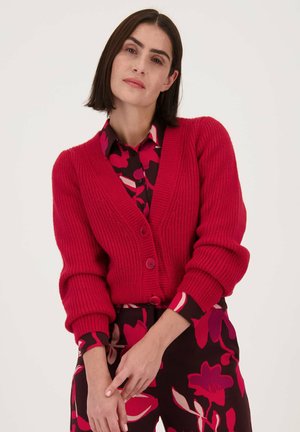 Cardigan rouge en tricot avec un col en V, présentant une texture côtelée et trois boutons. Porté sur un blouse à imprimé floral avec des tons roses et bordeaux.