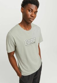 Junger Mann mit kurzen lockigen Haaren trägt ein hellgraues T-Shirt mit dem Text "CORE" und steht mit den Händen in den Taschen vor einem schlichten Hintergrund.