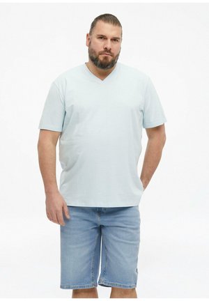 Mężczyzna z brodą w jasnoniebieskiej koszulce z dekoltem w serek i szortach jeansowych na jednolitym białym tle.