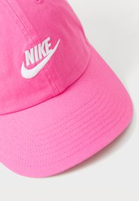 Rožinė medvilninė beisbolo kepurė su balta siuvinėtą „Nike“ logotipu. Iškraipytas vizorėlis su tekstūruota medžiaga. Paprastas dizainas, jokių papildomų akcentų.